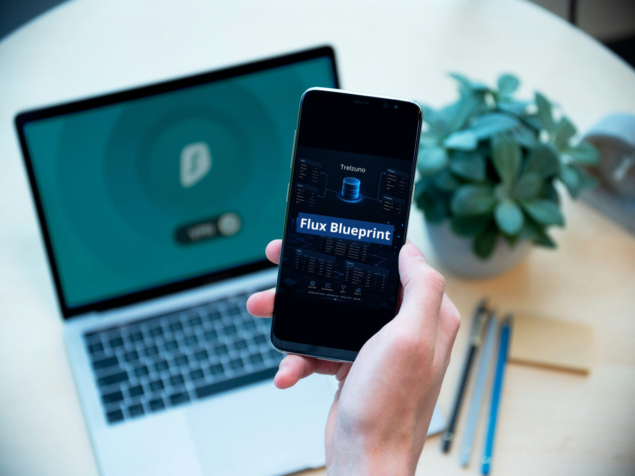 Person holding a smartphone with 'Flux Blueprint' app on a desk with laptop and plant